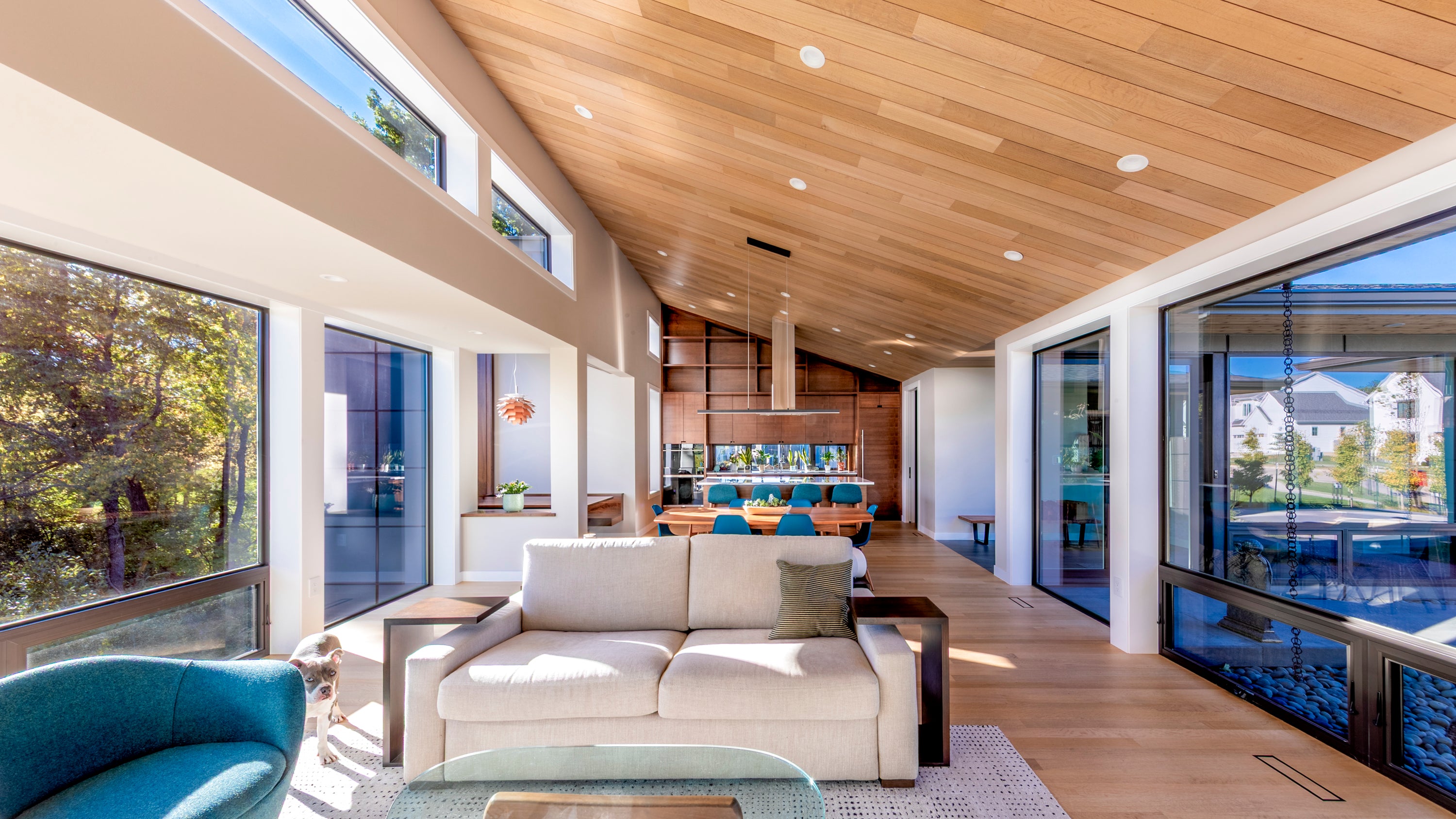 Modern living room with large windows, wooden ceiling, and kitchen in the background.