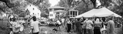 Black and white photo of a community gathering with people, tents, and modern house addition in the background.