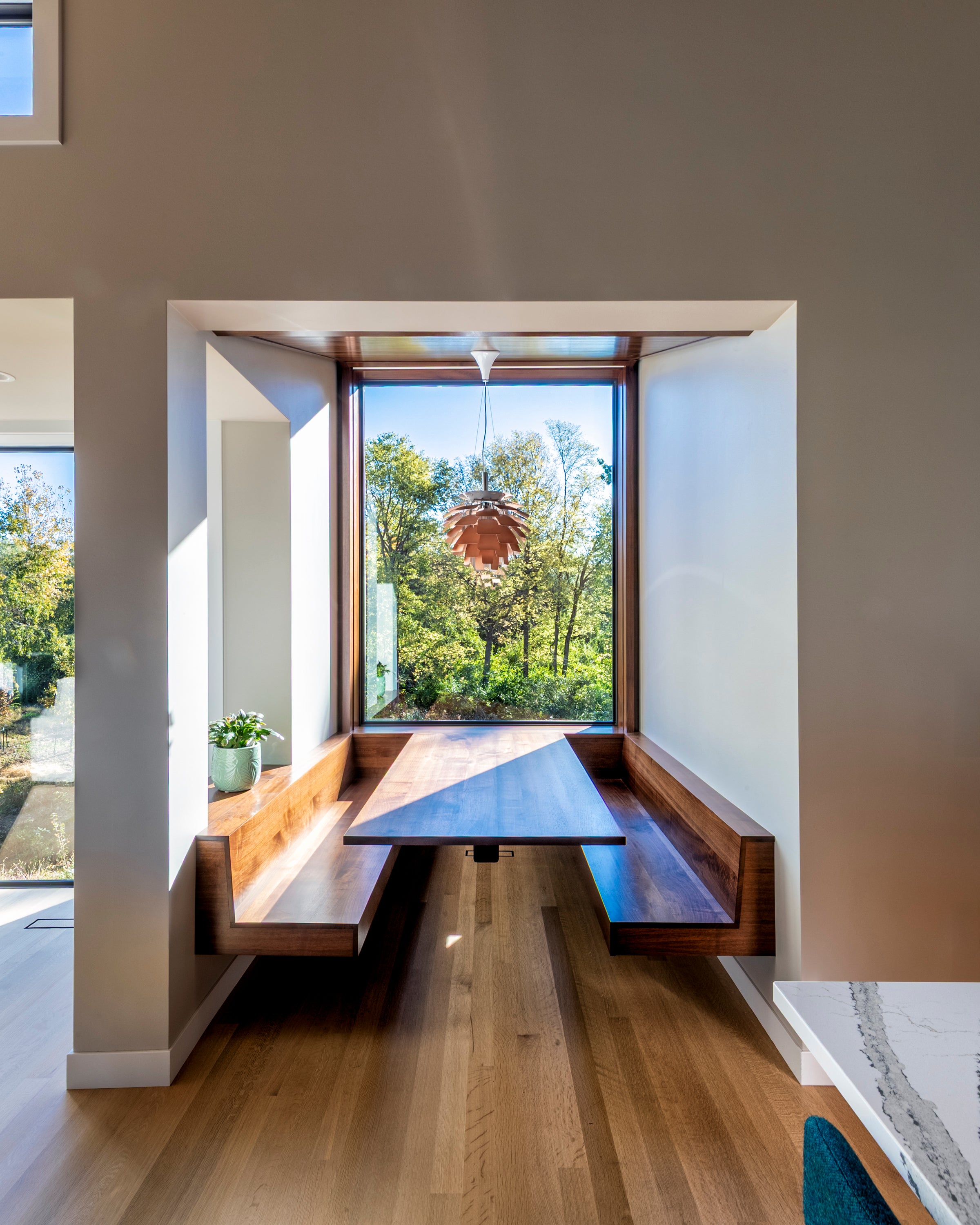 Modern interior with a wooden dining table and benches by large windows with a view of trees.