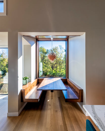 Modern interior with a wooden dining table and benches by large windows with a view of trees.